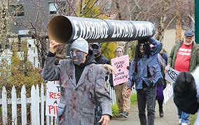 Photo shows protesters with mock section of pipeline, one dressed in gray, wearing a zombie mask.